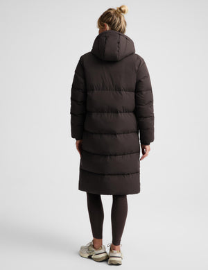 A woman viewed from behind wearing a long dark brown puffer coat, dark leggings, and sneakers against a light gray studio background.