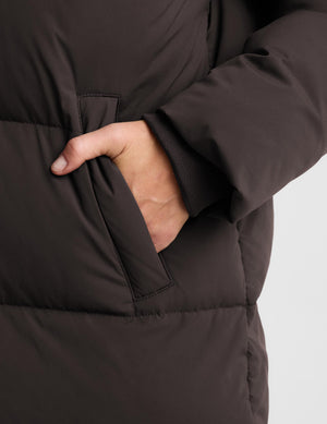 Close-up of a person wearing a dark brown puffer jacket, showing a hand reaching into a side pocket against a light gray background.