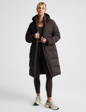 A woman modeling a long, dark brown puffer coat with a hood, worn over a matching dark brown activewear set and white sneakers, against a light gray studio background.