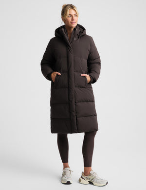A woman modeling a long, dark brown puffer coat with a hood, worn over dark leggings and white sneakers, against a light gray studio background.
