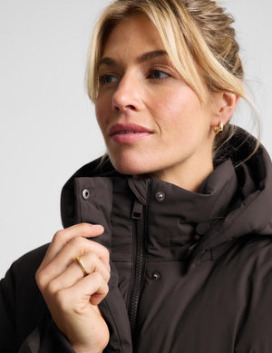 A close-up of a woman wearing a dark brown hooded jacket, pulling the collar up with one hand. She has blonde hair pulled back and is wearing a gold ring and earring against a light gray background.