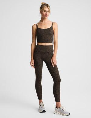 A woman modeling a matching dark brown and black speckled activewear set, consisting of a cropped tank top and high-waisted leggings, paired with light gray athletic sneakers, against a plain light gray background.