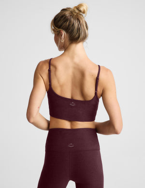 A woman modeling a dark burgundy sports bra and matching high-waisted leggings against a light gray studio background.