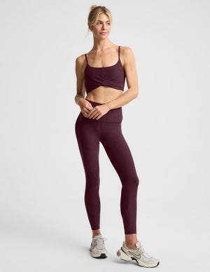 A woman modeling a dark burgundy sports bra and matching high-waisted leggings against a light gray studio background.