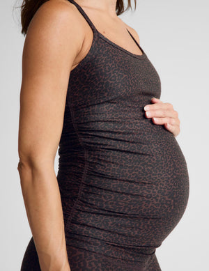 A pregnant woman wearing a dark brown and black leopard-print maternity tank top and matching leggings, standing against a light gray studio background.
