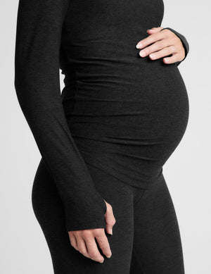 A pregnant woman wearing a black long-sleeve maternity top and matching leggings, standing against a light gray studio background.