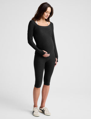 A pregnant woman wearing a black long-sleeve maternity top and matching leggings, standing against a light gray studio background.