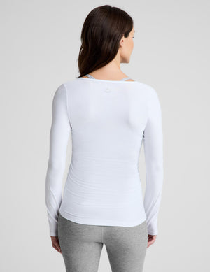 A pregnant woman wearing a white long-sleeve scoop-neck maternity top and gray leggings, standing against a light gray studio background.