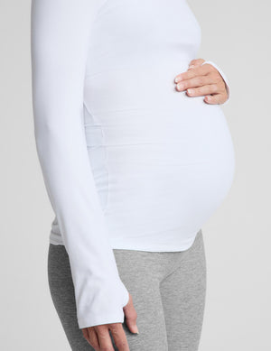 A pregnant woman wearing a white long-sleeve scoop-neck maternity top and gray leggings, standing against a light gray studio background.