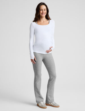 A pregnant woman wearing a white long-sleeve scoop-neck maternity top and gray leggings, standing against a light gray studio background.