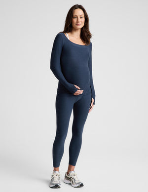 A pregnant woman modeling a navy blue long-sleeve maternity top and matching leggings set, paired with white and gray sneakers, against a light gray studio background.