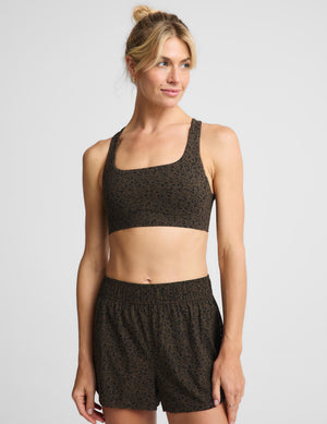 A woman modeling a matching dark brown and black leopard-print activewear set, featuring a sports bra and shorts, against a light gray studio background.