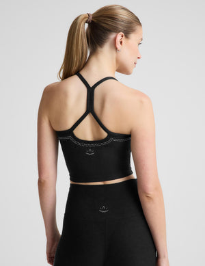 A woman wearing a black bedazzled crop tank with crisscross straps and a small white logo, paired with matching black leggings against a light gray studio background. 