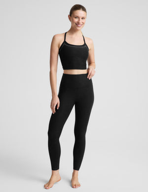 A woman wearing a black bedazzled crop tank with crisscross straps and a small white logo, paired with matching black leggings against a light gray studio background. 