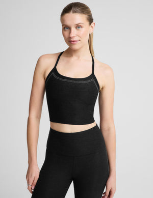 A woman wearing a black bedazzled crop tank with crisscross straps and a small white logo, paired with matching black leggings against a light gray studio background. 