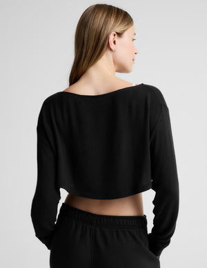 A woman shown from the back wearing a black long-sleeve cropped top with a wide boat neckline and black sweatpants against a light gray studio background.