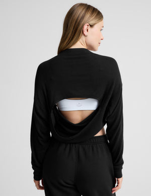A woman shown from the back wearing a black long-sleeve cropped sweatshirt with a draped open-back design, paired with black sweatpants and a white sports bra, against a light gray studio background.