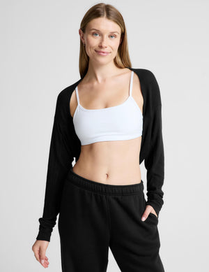 A woman modeling a white sports bra and black cardigan and sweatpants set against a light gray studio background.