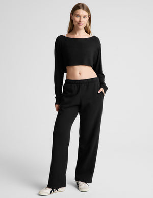 A woman modeling a black two-piece loungewear set consisting of a long-sleeve cropped top and matching wide-leg sweatpants, paired with white sneakers, against a light gray studio background.