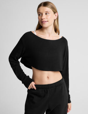 A woman modeling a black long-sleeve cropped top with a wide boat neckline, paired with black sweatpants, against a light gray studio background.