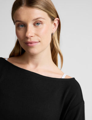 A close-up shot of a woman wearing a black off-the-shoulder knit top, showing her face, hair, and the black fabric texture against a light gray background.
