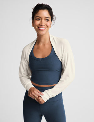 A woman modeling a matching activewear set consisting of a cream off-the-shoulder long-sleeve crop top and high-waisted navy blue leggings, paired with white sneakers, against a light gray studio background.