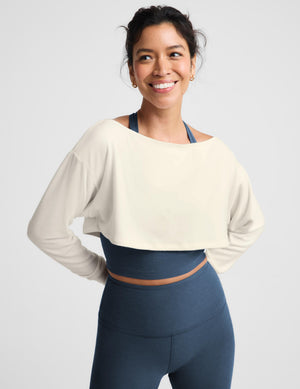 A woman modeling a matching activewear set consisting of a cream off-the-shoulder long-sleeve crop top and high-waisted navy blue leggings, paired with white sneakers, against a light gray studio background.