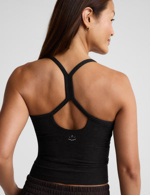 A woman shown from the back wearing a black knit camisole tank top and dark brown pants, standing against a light gray studio background.