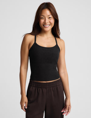 A smiling woman wearing a black knit camisole tank top and dark brown pants, standing against a light gray studio background.