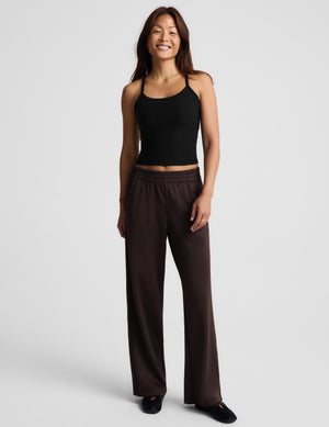 A smiling woman wearing a black knit camisole tank top and dark brown pants, standing against a light gray studio background.