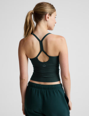A woman wearing a dark forest green ribbed tank top and matching green sweatpants, photographed against a neutral light gray studio background.