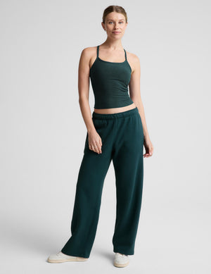 A woman wearing a dark forest green ribbed tank top and matching green sweatpants, photographed against a neutral light gray studio background.