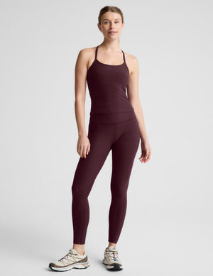 A woman modeling a dark plum-colored athletic tank top and matching leggings against a light gray studio background.