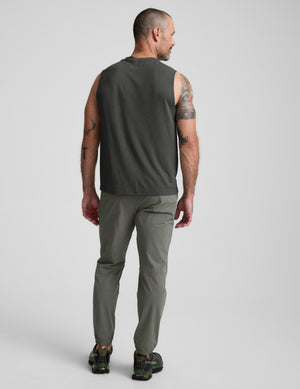 Man wearing a sleeveless dark gray shirt and green joggers on a light gray background. 
