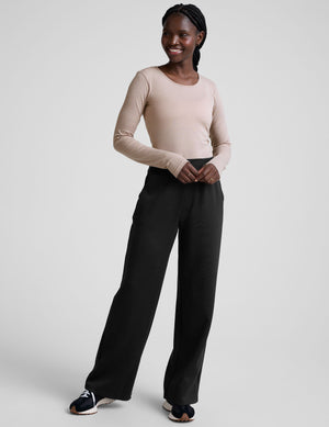 model wearing a beige long-sleeve crewneck top and black pants on a plain background. 