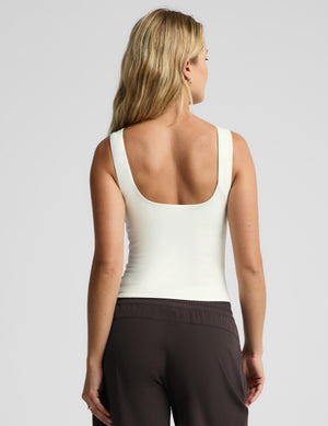 A blonde woman wearing a fitted sleeveless cream tank top and dark drawstring pants, photographed against a light gray studio background.