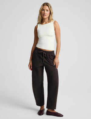 A blonde woman wearing a fitted sleeveless cream tank top and dark drawstring pants, photographed against a light gray studio background.