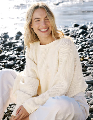 Woman in a white sweater sitting on a rocky beach. 